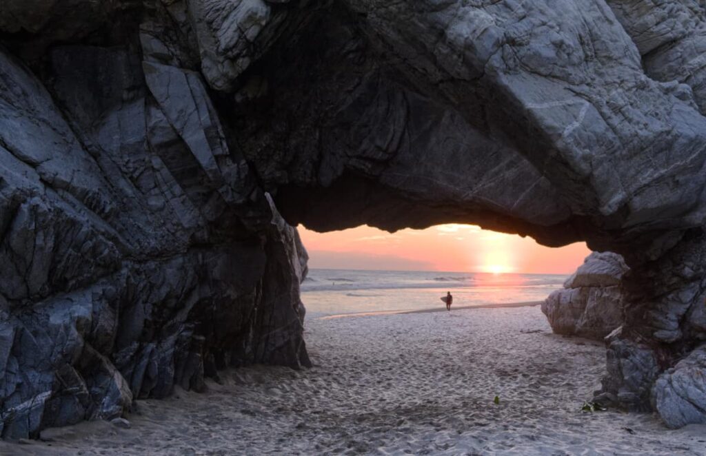 Framed within the dark rugged rock of Arco de Tiempo in Chacahua, a surfer holds a surfboard under their arm while walking along the beach. Behind them is a pink-orange sunset over the ocean.