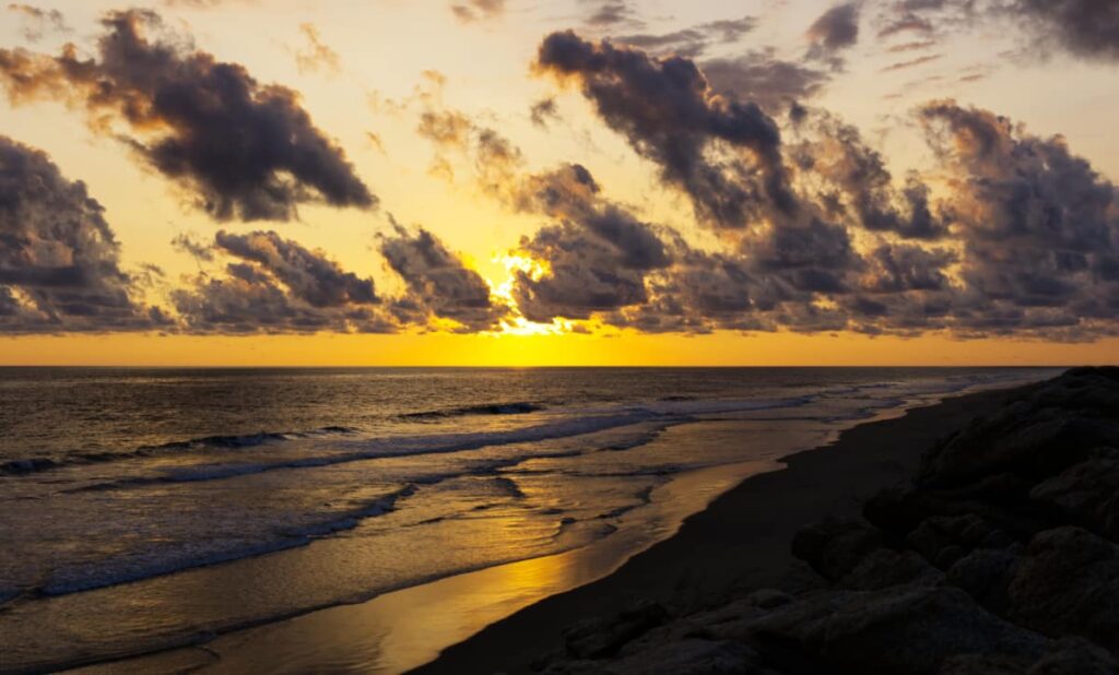 From atop Arco de Tiempo, clouds obscure the sunset over the ocean as small waves layer over the shore.