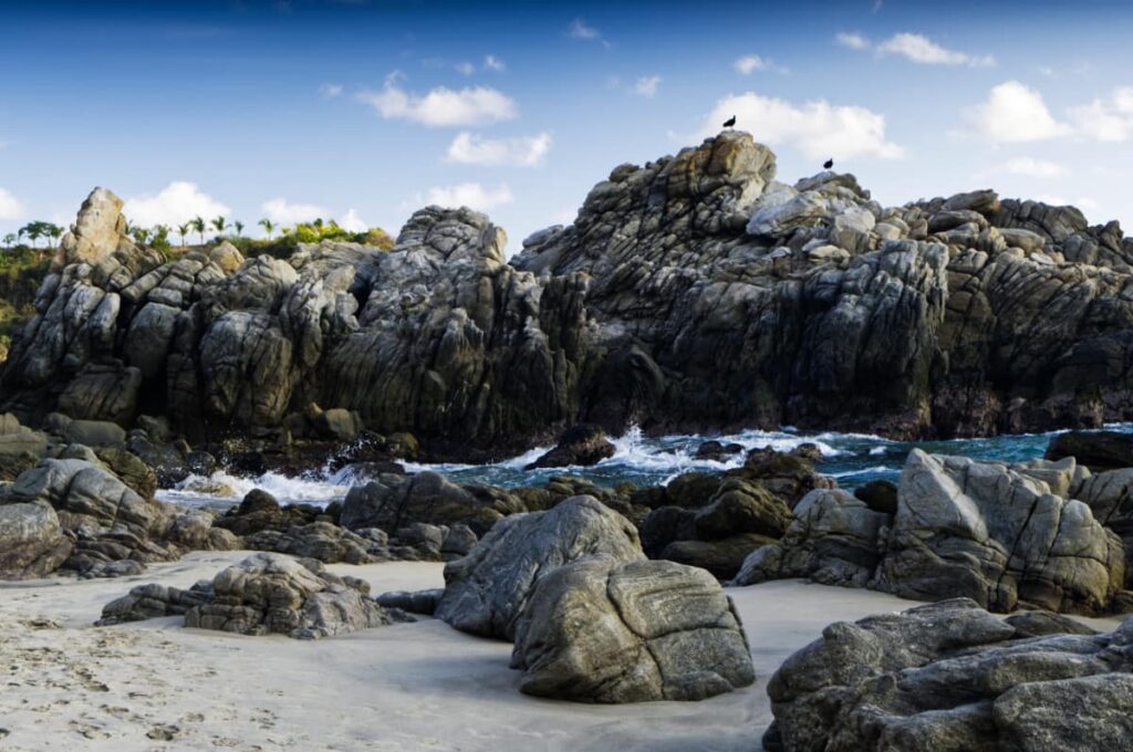 Large rocks with deep crevices sit in the sandy area between Bacocho and Playa Coral. Two vultures are perched on the tallest cliff face as the water flows in from the ocean.