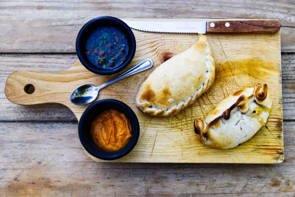 An overview shot of a wooden cutting board with two baked empanadas of different shapes, a small bowl of chimichurri, and another of hot sauce. There is also a steak knife and small spoon.