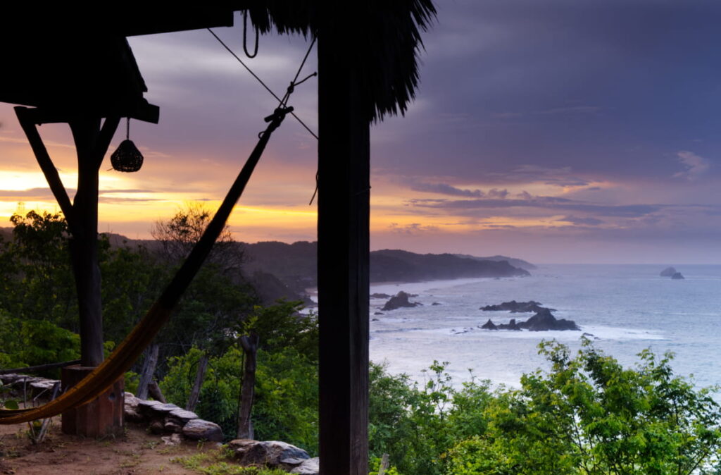 Looking past the hammock and cabin sitting area at Cabanas Balamjuyuc, a hostel in Mazunte, is the ocean view along the mountainous coastline. Above, the morning sky transitions from dark blue-purple clouds to bright orange and yellows.