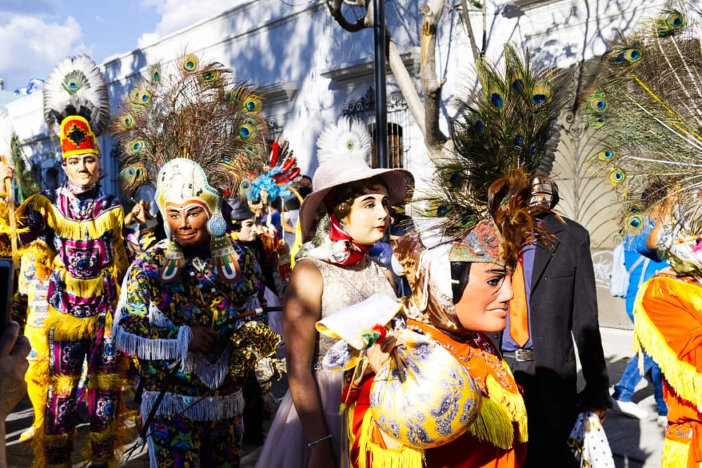 Dressed for the Carnaval Festival in Oaxaca, several people are lined up wearing masks that are painted like doll faces, both male and female. There are colorful costumes and peacock feathers.