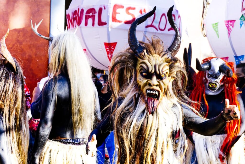 At the Oaxaca Carnaval, a man faces the camera wearing horns and a devil mask with the tongue sticking out. His body is painted black with motor oil and his hair is covered with a long flowing blonde animal hair. Behind him are others in similar costume.