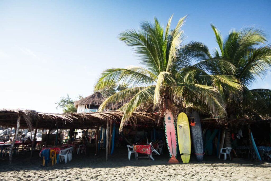 Pink, yellow, and grey surfboards lean against a palm tree near the surf spot in Chacahua. On the left are plastic tables and chairs under a palapa beachside restaurant.