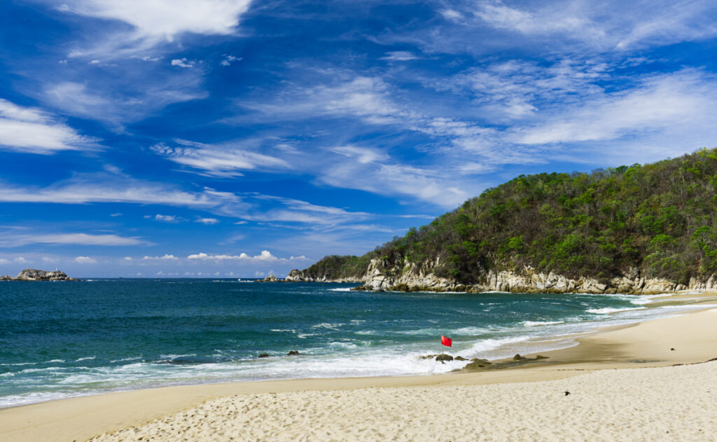A red flag anchored in the rocks on a beach in Huatulco indicates it is not safe for swimming. The bay has light golden sand, turquoise water, and a cliff with green trees.