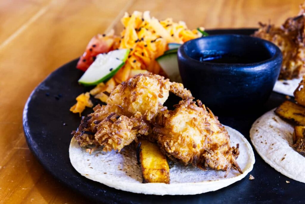 Fried coconut shrimp with sticks of fried plantain on a corn tortilla. A sauce sits in a black clay vessel in the center with a shredded carrot salad in the background.