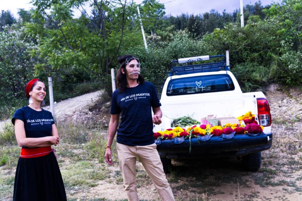 Carlos and his female associate wear Coyote Aventuras shirts that say Dia de los Muertos. Behind them is a white pickup truck, the tailgate down and with different flowers on top.