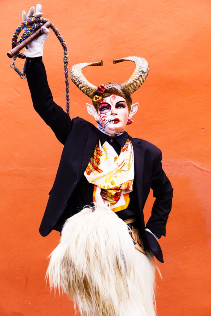 One of the devils of Santiago Juxtlahuaca poses in front of a bright orange wall. He is wearing a painted mask with pointed ears and animal horns. In his raised right hand is a colorful whipping rope. His clothes consist of a dark jacket, color scarf, and chaps made with cream colored animal fur.
