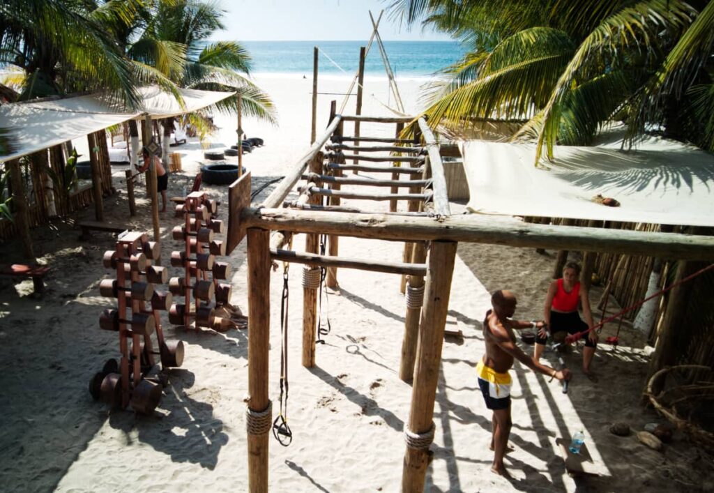 At this eco gym in Puerto Escondido, a shirtless man works out in the sand while a woman watches. Wooden dumbbells, monkey bars, and other gym equipment sit behind them.