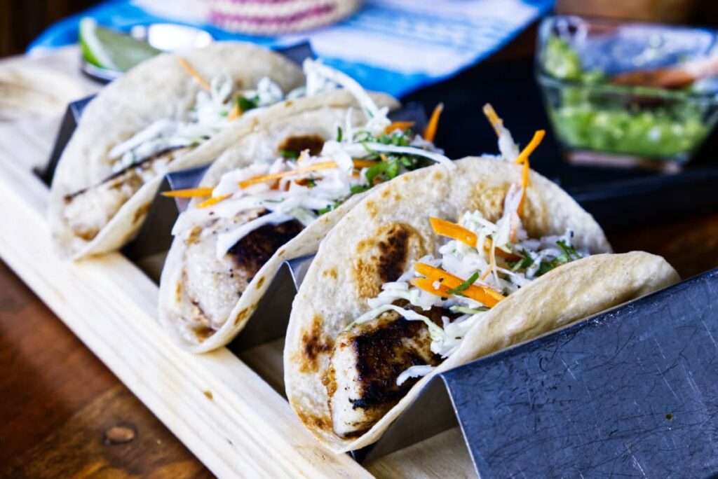 A metal taco stand holds three fish tacos loaded with shredded cabbage and carrots. In the background is a bright green bowl of tomatillo salsa.