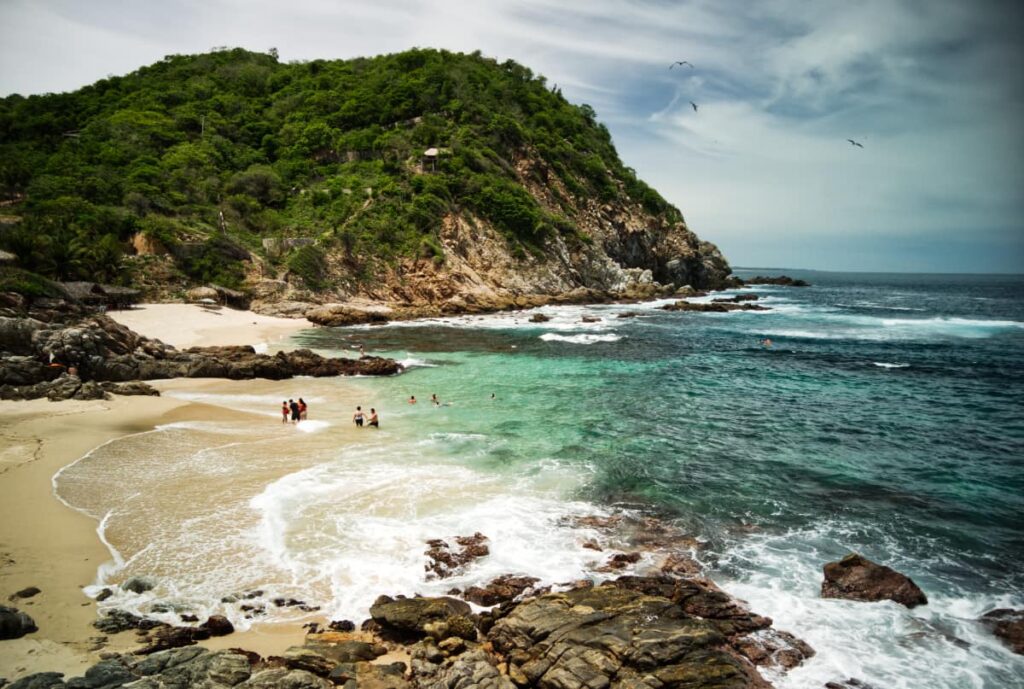 At Estacahuite Beach, Oaxaca people swim in a bay of turquoise water with golden sand and dramatic green cliffs behind them.