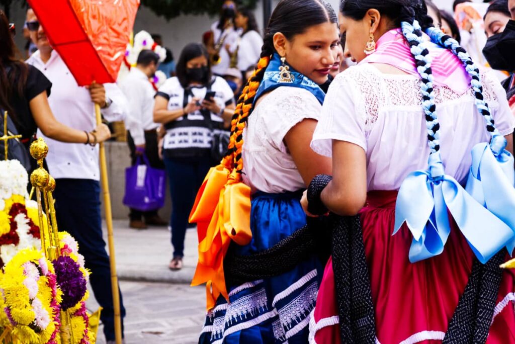 Young women dressed in traditional clothing talk as they wait for a parade during one of the biggest festivals in Oaxaca to begin. Their long hair is braided with colorful ribbons that hang down their back, tied in a bow.