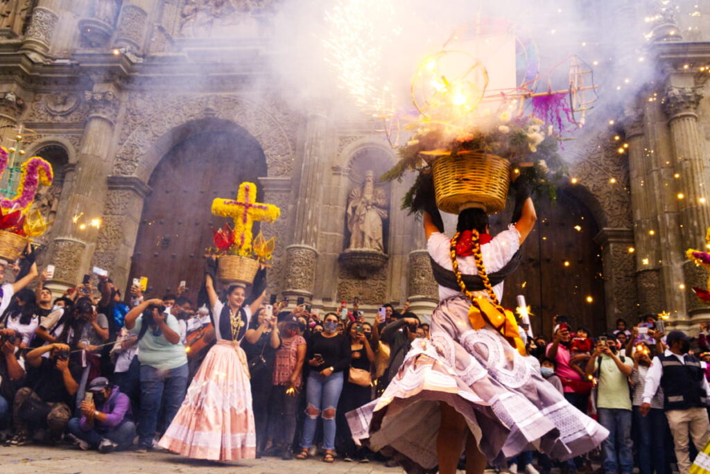 A woman dressed in traditional clothes dances at a Guelaguetza parade in front of the church. Two braids woven with bright yellow ribbon flow down her back. On top of her head is a basket of flowers and lit sparklers. On the left a women, also dressed in traditional clothes, holds a basket decorated with a large cross statue made of flowers. Much of the audience is taking pictures in the background.
