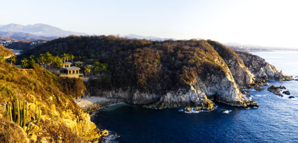 Sunrise illuminates the dramatic cliff face in Huatulco, along the coast of Oaxaca. In the bay is a a house and palms in front of a small beach with mountains in the background.