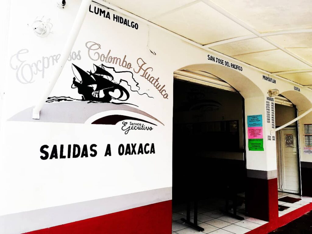 On the outside of the building for transportation from Huatulco to Oaxaca, hand painted letters and a boat graphic are painted on the wall. They say Expresos Colombo Huatulco and Salidas a Oaxaca.Above the archway entrances are the painted letters of towns they stop at, including Pluma Hidalgo and San Jose del Pacifico.
