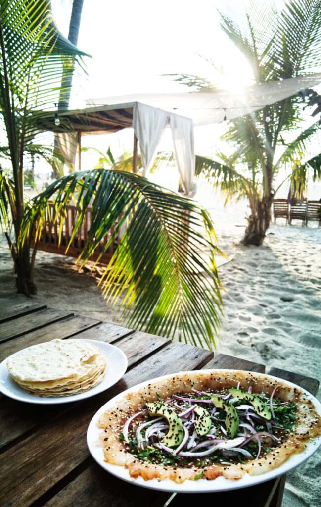 A plate of fresh sashimi fish topped with slices of red onion and avocado sits on a table at La Mariinera Restaurant in Zicatela. In the background is a sunbed in the sand surrounded by short palms with the sun streaming through the leaves.