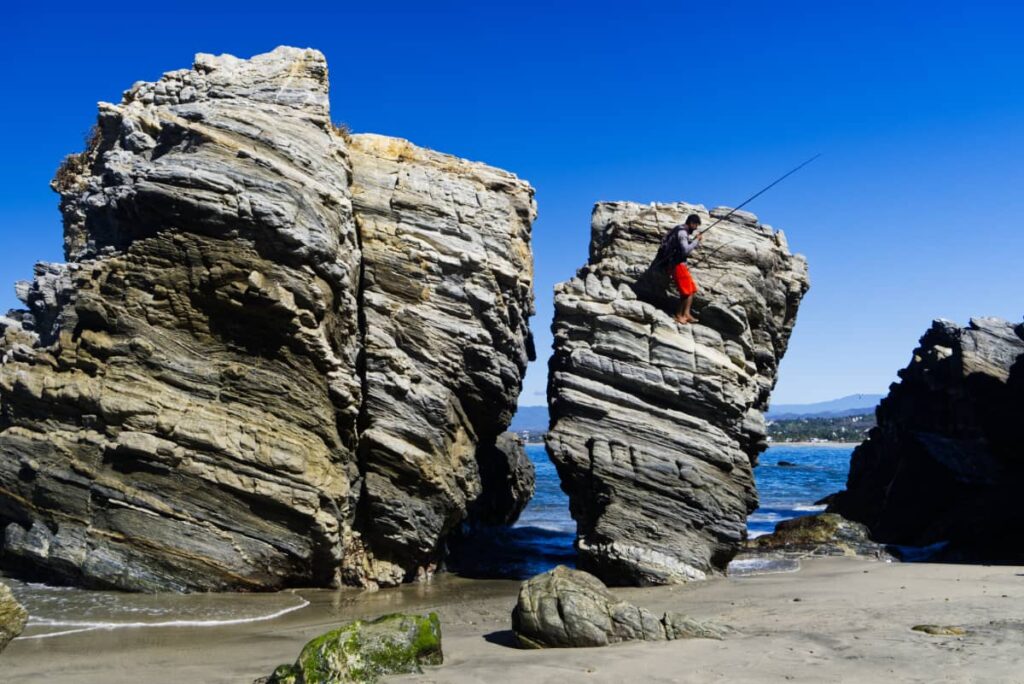 A fisherman in red shorts climbs down a large marbled rock with a fishing rod in his hand. These large rock formations near the ocean are the highlight of this hike, one of the best things to do in Puerto Escondido.