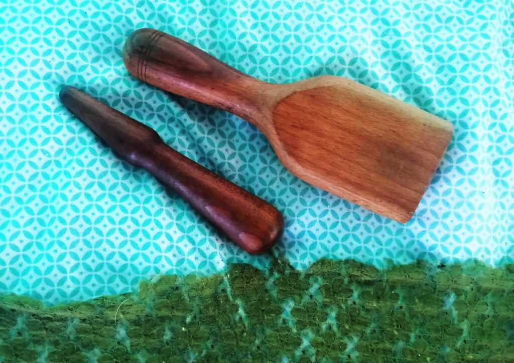 Two wooden tools that are used for massage lay on a colorful teal sheet. One resembles a chisel with a wider point. The other looks like a fully wooden paint brush.