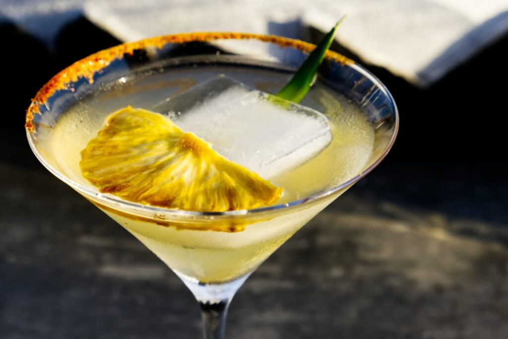 A refreshing cocktail in a martini glass with a partial rim of red spicy salt, a slice of pineapple, large ice cube, and greenery.