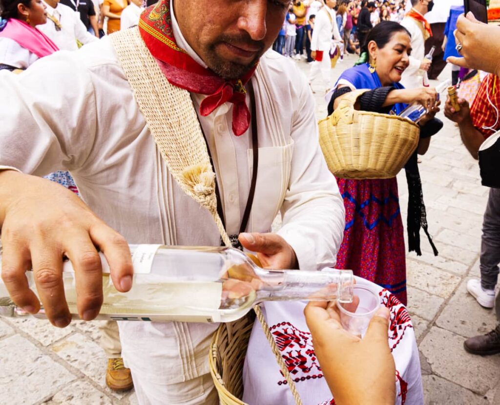 During the Guelaguetza Festival in Oaxaca, a man pours a spectator a shot of mezcal. In the background a woman holding a basket pours a different spectator a shot of mezcal.