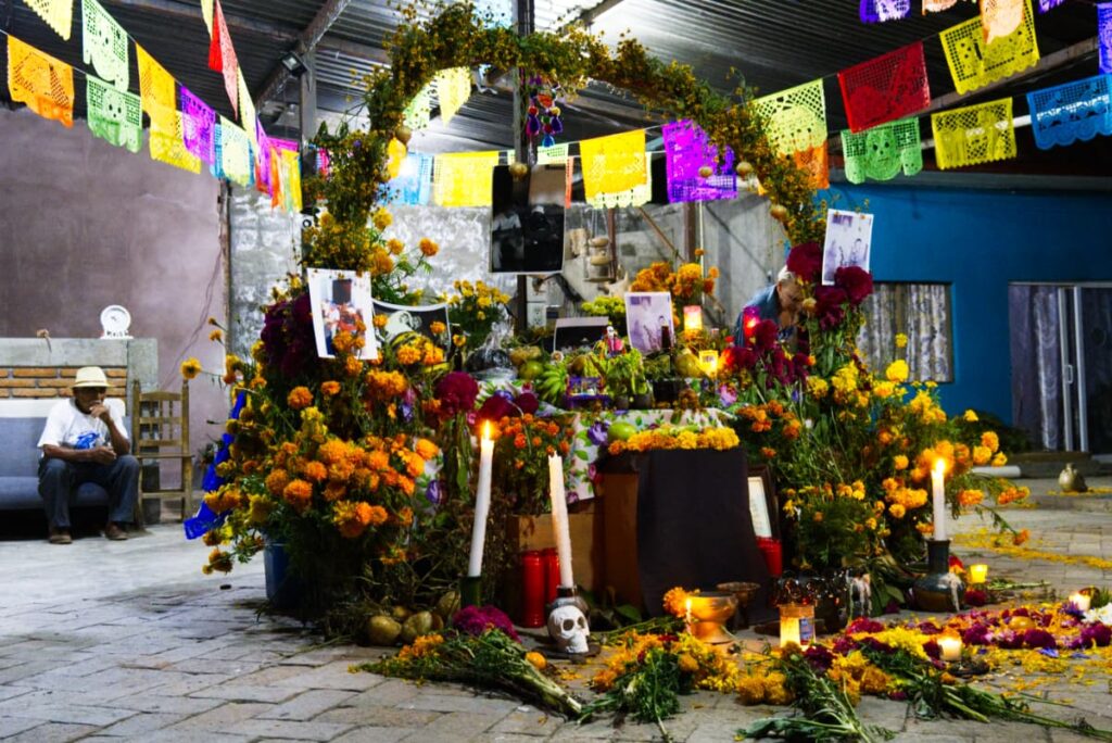 Tall candles light another beautiful muertos altar. There are lots of flowers, pictures of the deceased and a green arch hovering over it all. Colorful Mexican paper flags hang above and a man sits in contemplation on the left.