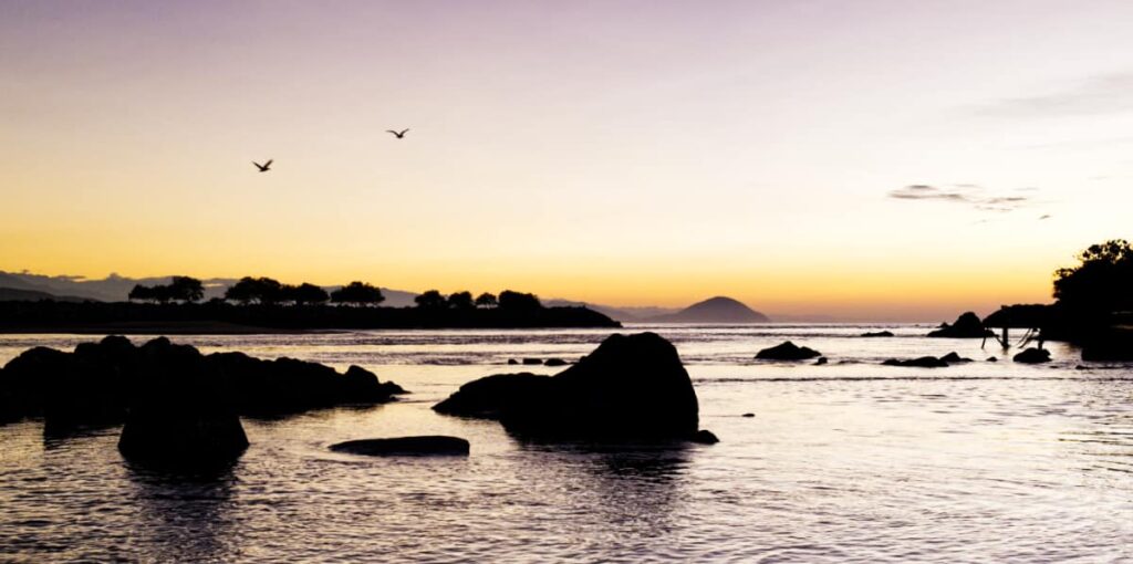 Two birds fly above the Chacahua lagoon. The morning light silhouettes the rocks sitting in the shallow water and reflects off the surface.