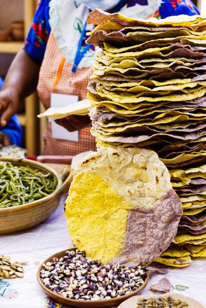 At the Oaxaca Festival of Traditional Chefs, a stack of tostadas (hard tortillas) of mixed colors sits on a table. In front is a bowl of purple corn, showing the various corn used for tortillas in Oaxaca.