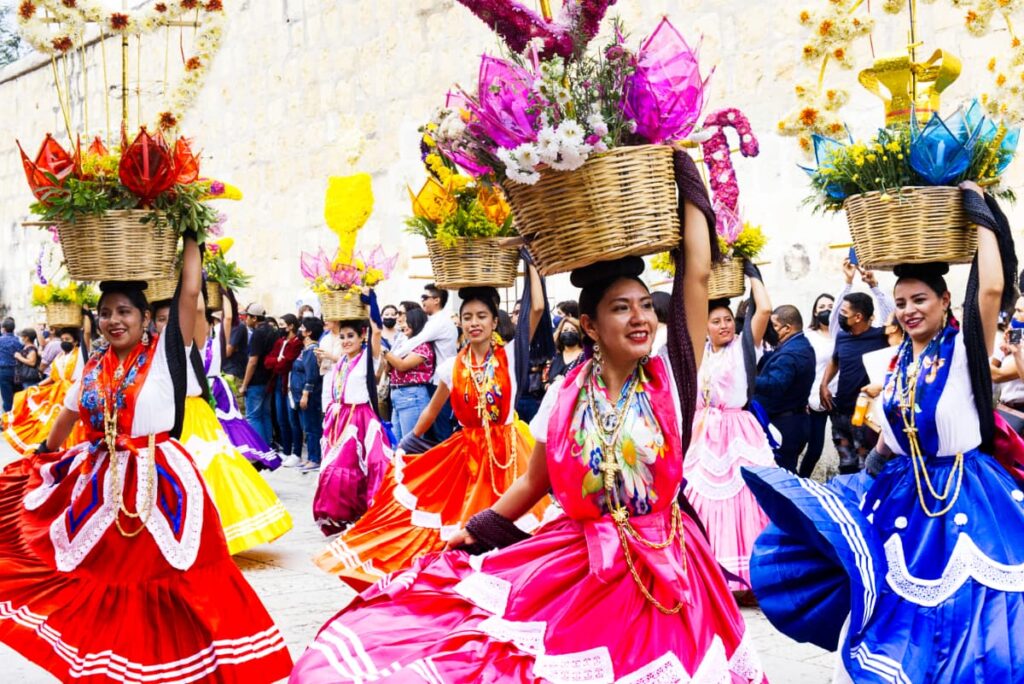 During the Guelaguetza Festival in Oaxaca, a man pours a spectator a shot of mezcal. In the background a woman holding a basket pours a different spectator a shot of mezcal.
