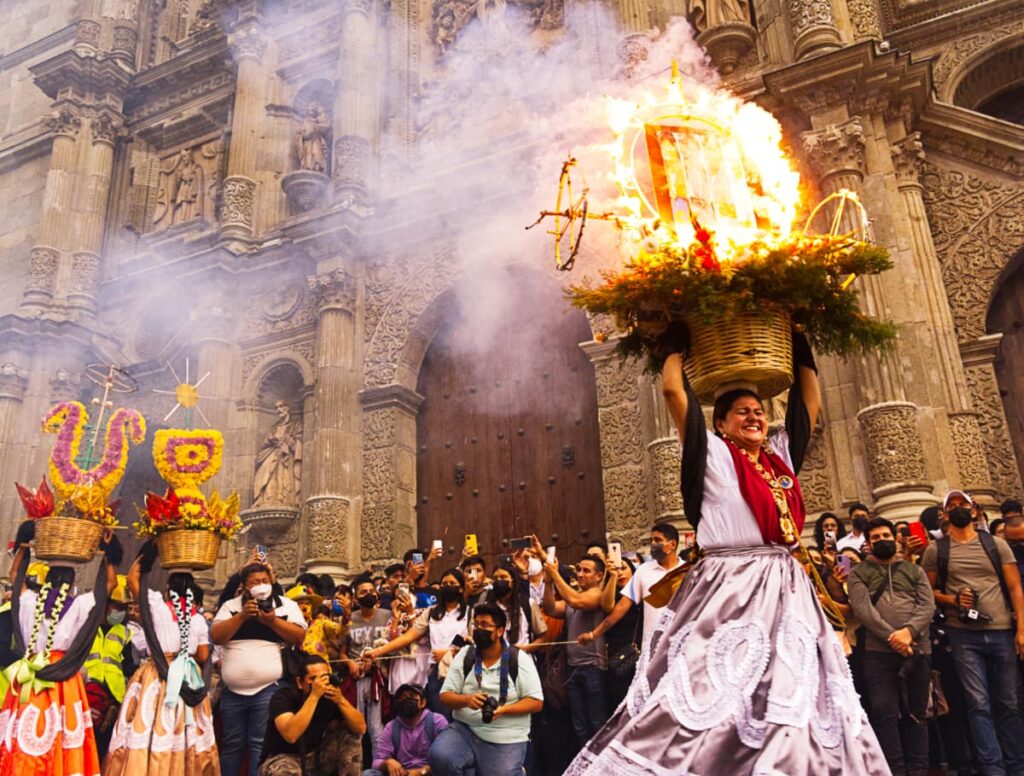 A woman with a large smile across her face dances at the Guelaguetza Parade in Oaxaca. She is dressed in traditional clothes and is holding a basket on top of her head that is filled with flowers and a circular structure that is laced with sparklers. The main structure is completely lit, with smoke billowing off to the side. In the background, the audience watches in amazement and take pictures.
