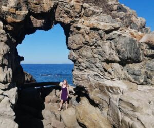 Julien, the creator of Oaxaca Travel Tips, stands in an rock arch that frames the ocean in La Punta, Oaxaca. She's wearing a purple dress and has long blonde hair.