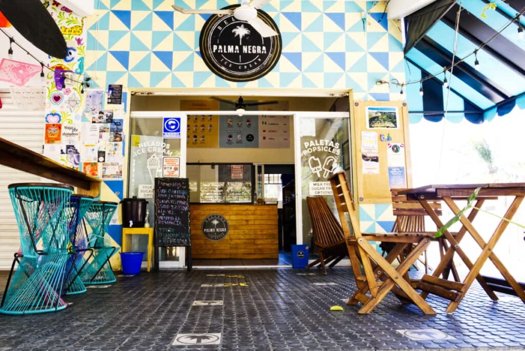 Outside of Palma Negra, the best ice cream in Puerto Escondido, are several tables and chairs. On the door signs feature words and graphics of ice cream and popsicles.