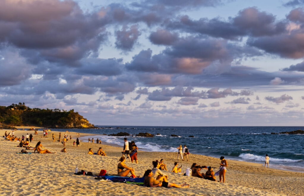 Groups of people walk and lounge on Playa Bacocho while the setting sun casts a golden hue across the beach. This is one of the best places to watch the sunset in Puerto Escondido, Mexico.