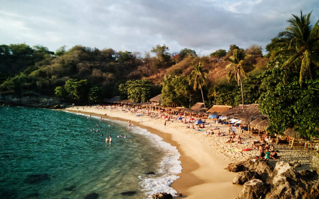 Golden sand and turquoise waters sweep along the bay at Playa Carrizalillo,  a great relief from the hot and humid weather in Puerto Escondido. People lounge in the sand in front of the palapa shaded restaurants while others swim in the ocean.