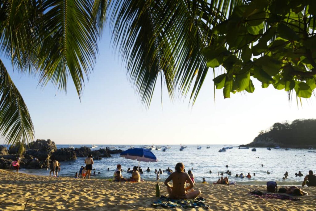 At one of the best Puerto Escondido beaches, a woman sits in the sand facing the ocean. Others also lounge and enjoy swimming in the water of this calm bay.