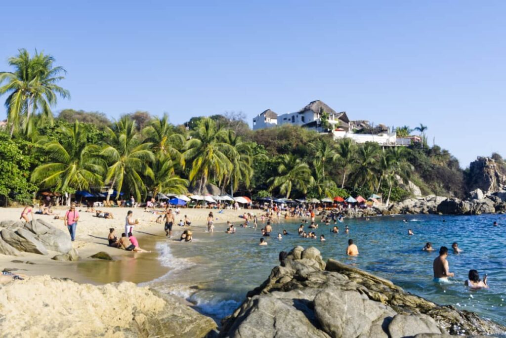 Just beyond the view of the rocks, lots of people swim and walk along the beach at Playa Manzanillo in Puerto Escondido. Short palm trees line the back cliff in which a large hotel is situated.