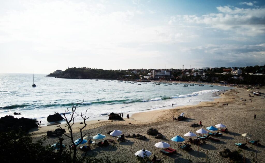 An overview of Playa Mainero, Puerto Escondido. Umbrellas and lounge chairs are lined up in the sand, a sailboat is anchored in the bay, and a few people walk around. In the background is low skyline of Puerto Escondido.