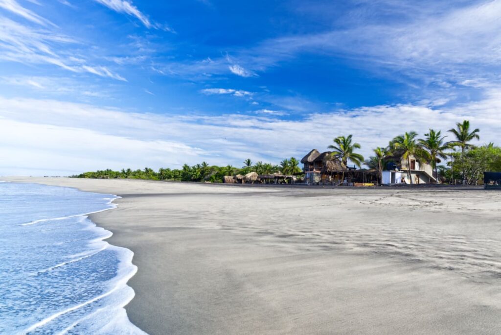 A small wave washes over the sand at Playa Ventanilla where palm trees line the beach among a few houses.