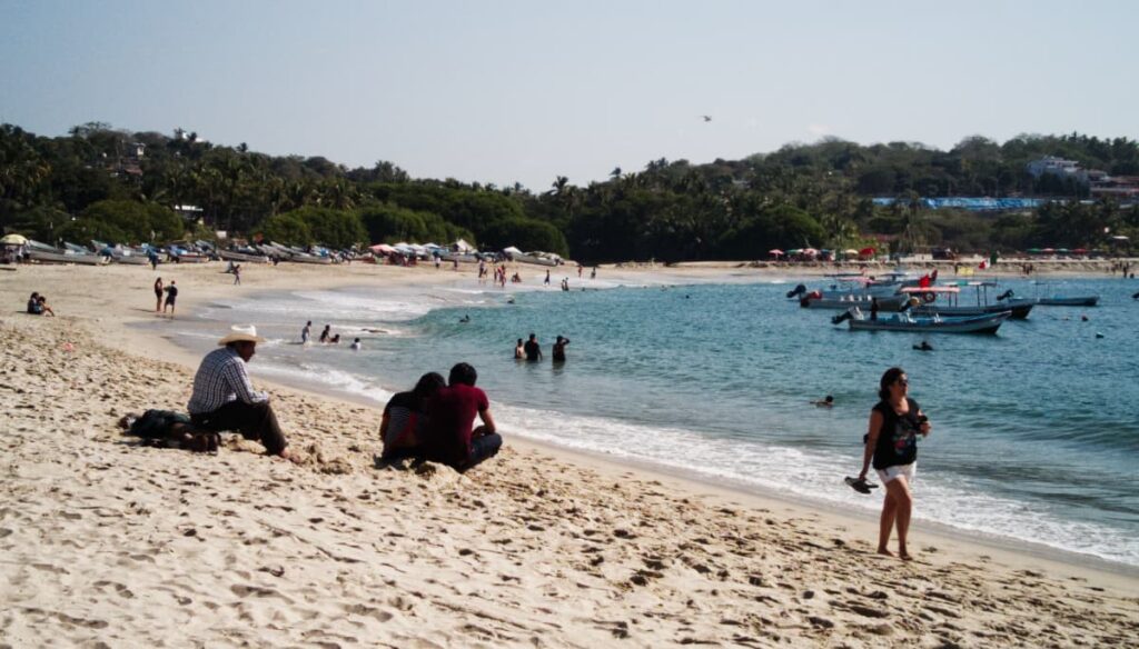 At Bahia Principal, the central beach in Puerto Escondido, a woman strolls along the shoreline with sandals in her hand. Several people including a couple and a man in a cowboy hat sit in the sand while others play in the ocean. Several boats are also anchored in the bay.