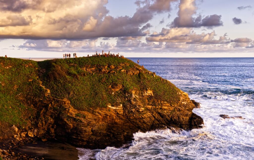 About 30 people gather on the top of the cliff at Punta Cometa to watch the sunset while waves roll into the sandy cove below.