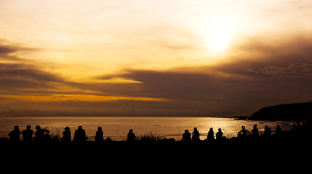 Silhouetted against the setting sun, about 15 people sit in a line along the edge of the cliff to watch the sunset at Punta Cometa.