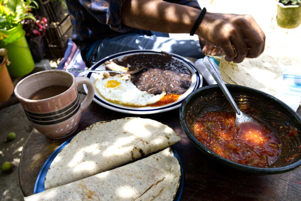 A man pulls apart a tortilla in a table view shot. On the table is a plate of beans with sunny side up eggs, a large bowl of red salsa, a cup of hot chocolate, and a plate filled with two quesadillas.
