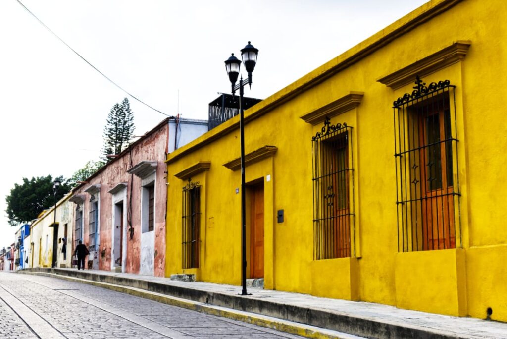 Colorful buildings line an empty street. Safety in Oaxaca can be a concern when they are this quiet. In the foreground is a house painted a bright mustard yellow.