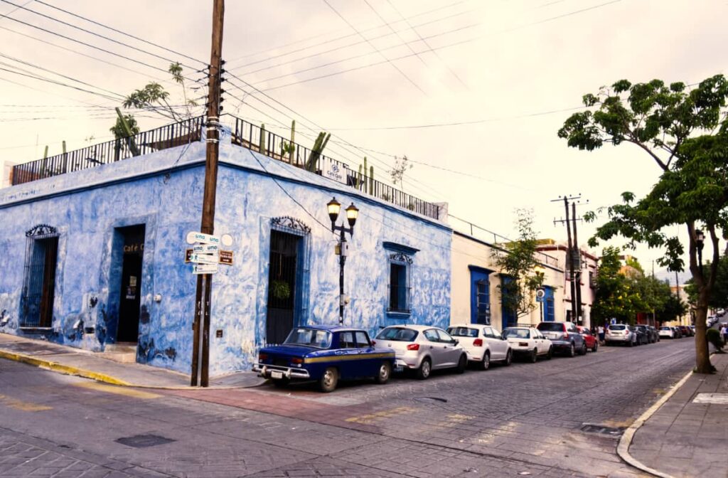 In front of a blue corner building with a rooftop, cars are parked on the street in Oaxaca.