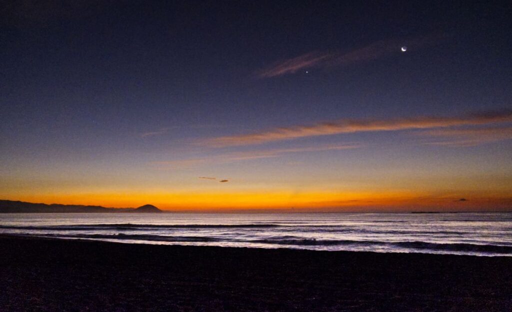 Intense hues of orange fill the sky above the ocean in Chacahua. There are small mountains in the distance and a crescent moon in the sky.