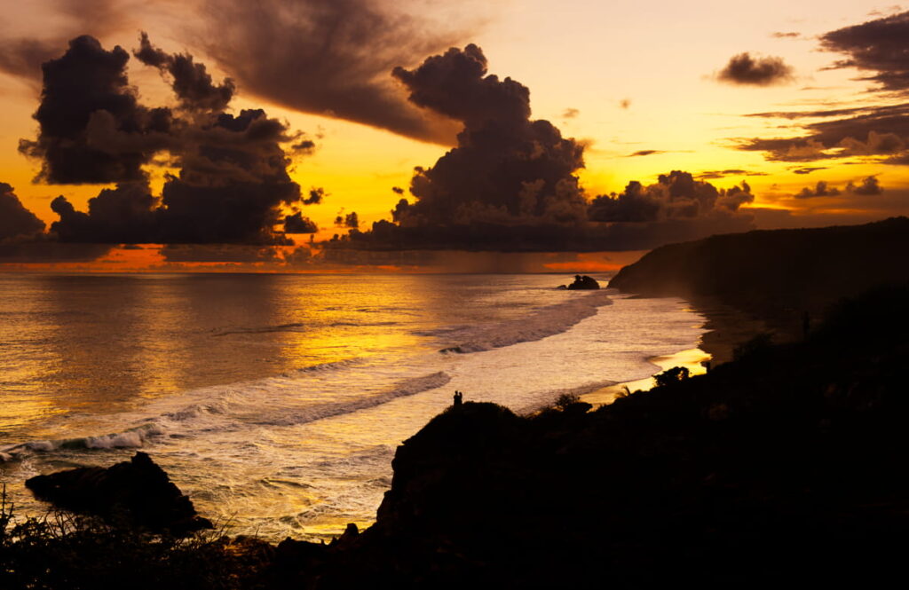 From afar, two people stand on the hill in front of the ocean and sunset at Punta Cometa, Oaxaca.