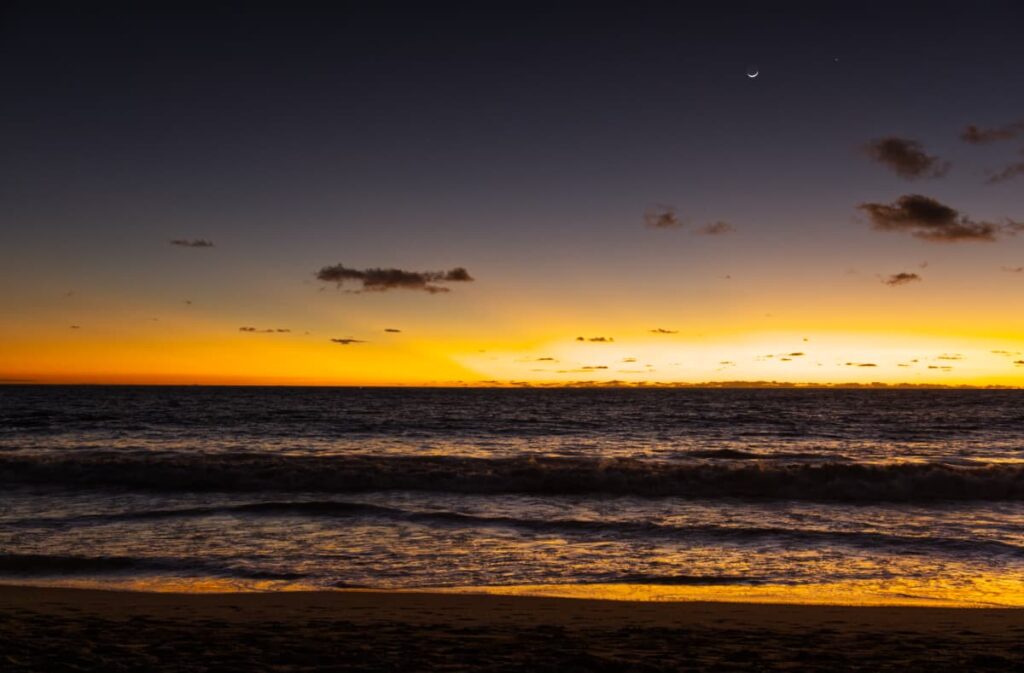 A crescent moon hangs above the ocean as the last colors of the sunset at Zicatela Beach fade away. As the waves recede from the shoreline, a golden hue is cast in the wet sand.