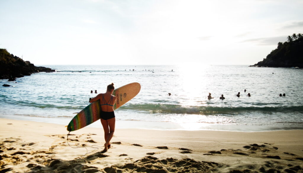 A girl walks towards the ocean with a colorful striped surfboard under her arm. Surf lessons are one of the top things to do in Puerto Escondido, Mexico.