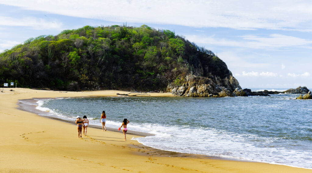 Four women wearing bikinis walk along one of the best beaches in Huatulco at Tangolunda Bay. The bay curves around to a large cliff that is covered with green trees.