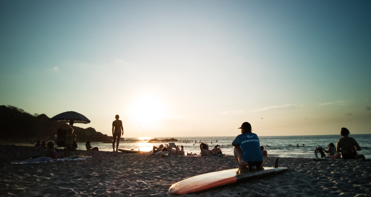 A woman squats on her surfboard which is laying in the sand at La Punta Beach. Others also sit and stand on the beach to watch the sunset.