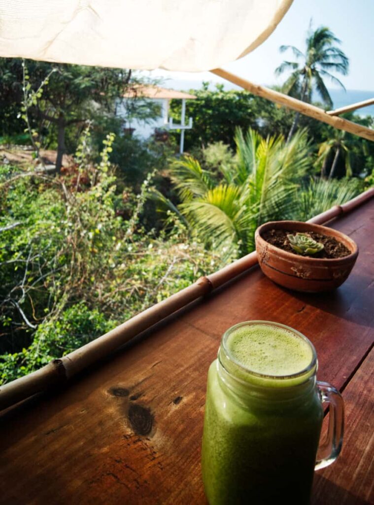 A jar mug is filled with green juice from Tistal Restaurant in Zicatela. In the background is an ocean view and palm tree.